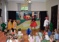 Na tle dekoracji stoi czworo dzieci przebranych w różne stroje oraz ubrana na czerwono kobieta. Przed nimi siedzą dzieci i oglądają przedstawienie.