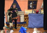 Na tle dekoracji stoi mężczyzna i dziecko przebrani w stroje leśniczych. Obok stoi łóżko, w którym znajduje się maskotka wilka. Przed występującymi siedzą dzieci i oglądają przedstawienie.