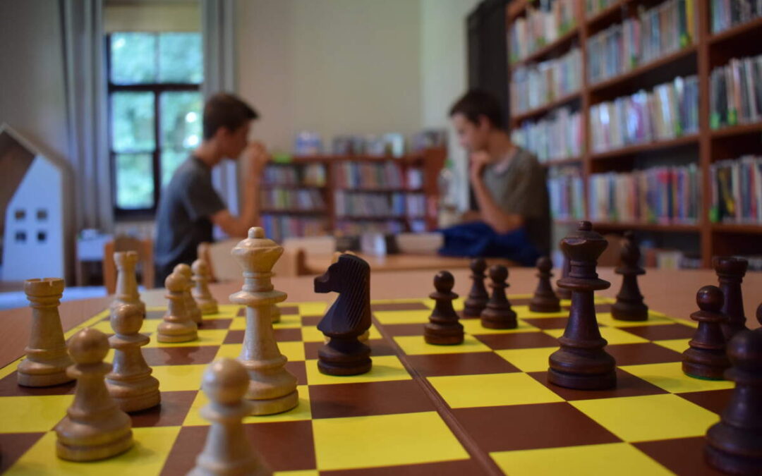 Na pierwszym planie plansza z rozstawionymi figurami szachowymi. W tle dwóch chłopców siedzi na przeciw siebie.