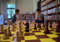 Na pierwszym planie plansza z rozstawionymi figurami szachowymi. W tle dwóch chłopców siedzi na przeciw siebie.