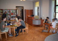 Widok całej sali. przy stołach siedzą dzieci i grają w szachy. W tle widać biurko i półki z książkami.